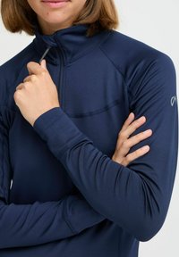 Tengerészkék, zipzáros, hosszú ujjú felső, sima textúrával, kontrasztos varrással, testhez álló fazonban, és egy kis logóval a váll közelében.