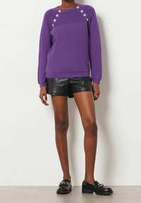 Paarse gebreide trui met geribbeld patroon, voorzien van zes decoratieve knopen op de schouders, gecombineerd met zwarte leren shorts en stevige zwarte schoenen.