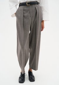 Grijze herringbone broek met plooien, gecombineerd met een zwarte riem met een gouden gesp, en zwarte veterschoenen. Slim fit.