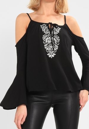 Vrouw draagt een zwarte cold-shoulder top met witte bloemenborduursel en wijde mouwen, gecombineerd met zwarte leren broek.