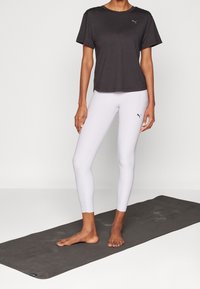 Svart kortärmad sport-T-shirt tillsammans med vita leggings, båda med Puma-logotypen. Barfota på en grå matta med textur.