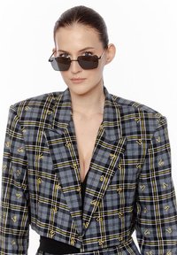 Kobieta z gładko zaczesanymi włosami, nosząca kanciaste ciemne okulary przeciwsłoneczne oraz skórzaną szaro-skalową marynarkę w kratę z żółtymi akcentami na czarnej bluzce.