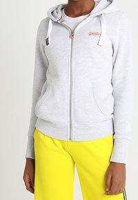 Felpa zip grigia chiara con cappuccio regolabile, tasche frontali e un piccolo logo ricamato. Abbinata a pantaloni gialli brillanti.