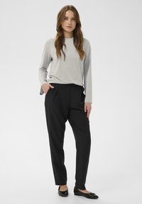 Sieviete stāv, ar roku kabatā, ģērbusies gaiši pelēkā garo piedurknu blūzē, melnās šaurās biksēs un melnās kurpēs pret vienkāršu fona.