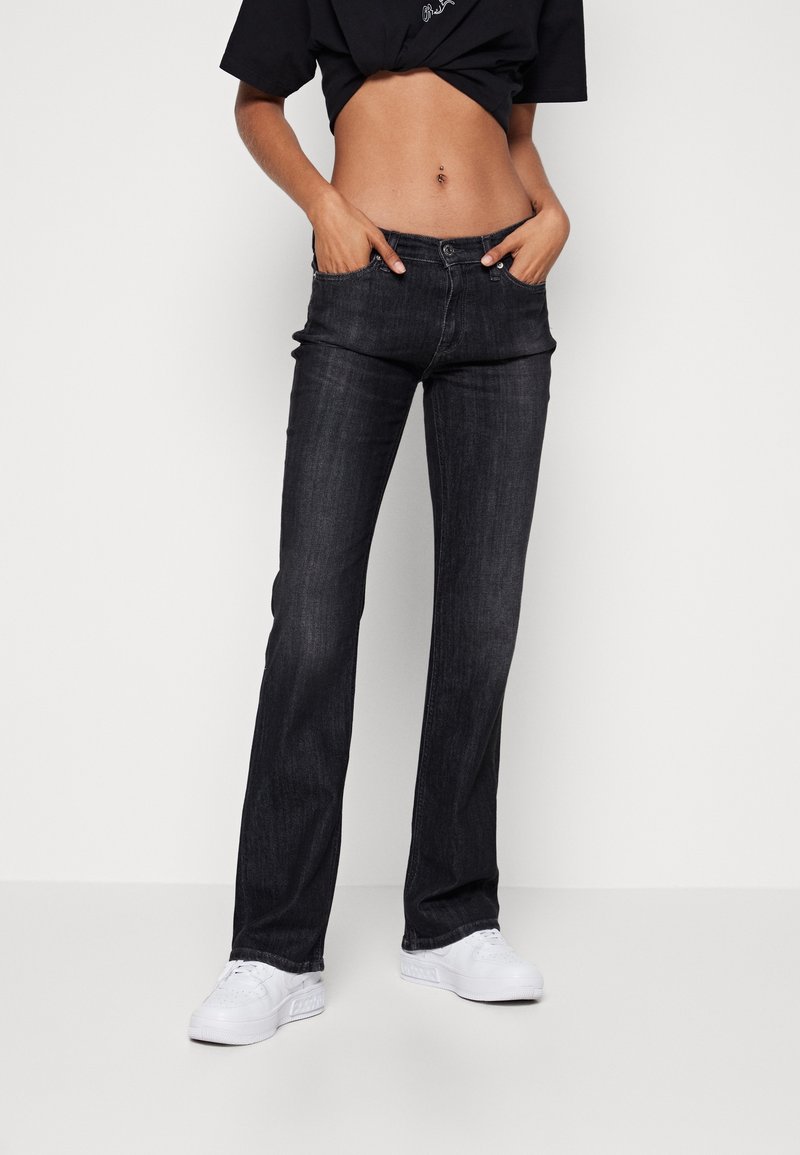 Personne portant un jean noir droit avec les mains dans les poches, des baskets blanches et un t-shirt noir court montrant un nombril percé.