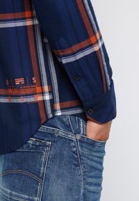 Mörkblå rutad skjorta med orange och vita ränder, två svarta knappar på ärmsluten, kombinerad med blå denimjeans med fickdetalj.