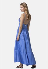 Abito lungo blu con gonna a strati, spalline sottili e schiena bassa. Realizzato in tessuto morbido con una vestibilità comoda. Sandali neri indossati ai piedi.