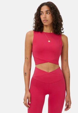 Vrouw met krullend donker haar die een mouwloos rood crop top en rode leggings met hoge taille draagt, staand tegen een effen lichte achtergrond.