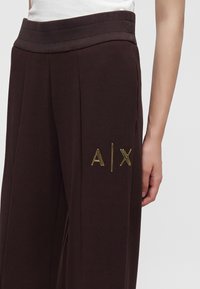 Pantalon large marron foncé avec une ceinture élastique et un logo doré A|X sur la cuisse droite, porté avec un haut blanc.