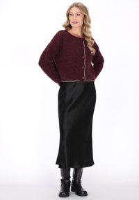 Kvinde med bølget blond hår iført en bourgogne knapstrikket sweater, sort lang satin nederdel og sorte læder ankelstøvler.