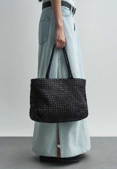 Μαύρη πλεκτή τσάντα tote κρατούμενη με το ένα χέρι, με πλεγμένα χερούλια, ορθογώνιο σχήμα και υφή στην επιφάνειά της. Τζιν φούστα που φτάνει μέχρι τους αστραγάλους.