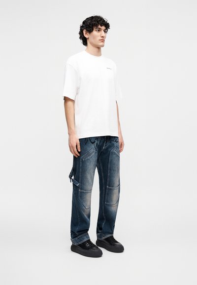Νεαρός με σγουρά μαλλιά στέκεται φορώντας λευκό φαρδύ t-shirt, άνετο μπλε τζιν παντελόνι με τσέπες και μαύρα αθλητικά παπούτσια σε απλό φόντο.