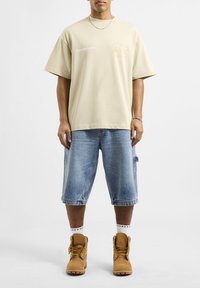 Beżowy, oversize T-shirt z logo, połączony z jasnoniebieskimi szortami dżinsowymi i beżowymi butami. Krótkie rękawy, luźny krój.