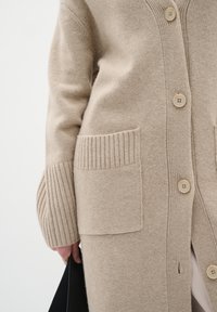 Beige stickad cardigan med ribbade muddar och två frampockets. Har runda knappar och en mjuk textur, vilket ger mångsidighet vid användning.