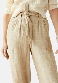 Beżowe lniane spodnie z fałdami z przodu i związanym paskiem z tkaniny noszone z kremowym swetrem dzianinowym.