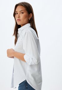 Camicia a righe blu chiaro con colletto button-down, maniche lunghe e decorazioni di perline sul davanti. Indossata con jeans in denim scuro.