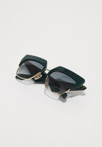 Gafas de sol verdes de gran tamaño con un acabado brillante, montura cuadrada, lentes de gradiente oscuro y acentos de metal dorado en las patillas.