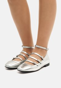 Ballerines métalliques argentées avec plusieurs fines lanières à la cheville et un design à bout pointu. Texture lisse et petit talon plat.