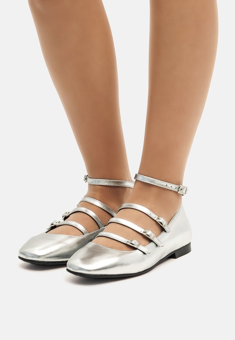 Ballerines métalliques argentées avec plusieurs fines lanières à la cheville et un design à bout pointu. Texture lisse et petit talon plat.