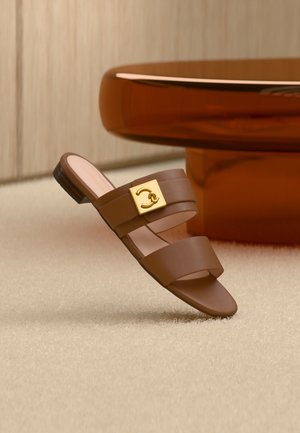 Sandale slide en cuir marron avec double brides, détail boucle carrée dorée, présentée sur un tapis beige près d'un pied de table rond marron.