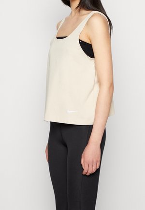 Kobieta ubrana w beżowy, bezrękawnik Nike na czarny sportowy stanik i czarne legginsy, stojąca bokiem na jednolitym tle.