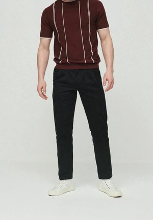 Homme portant une chemise en maille à manches courtes bordeaux avec des rayures verticales blanches, un pantalon noir et des baskets blanches, debout devant un fond uni.