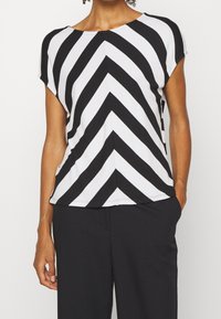 Frau trägt ein schwarz-weißes Chevron-streifen Kurzarmoberteil mit schwarzen Hosen, eine Hand in der Tasche, auf Hals und Taille gekürzt.