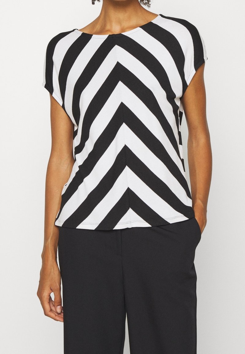 Frau trägt ein schwarz-weißes Chevron-streifen Kurzarmoberteil mit schwarzen Hosen, eine Hand in der Tasche, auf Hals und Taille gekürzt.