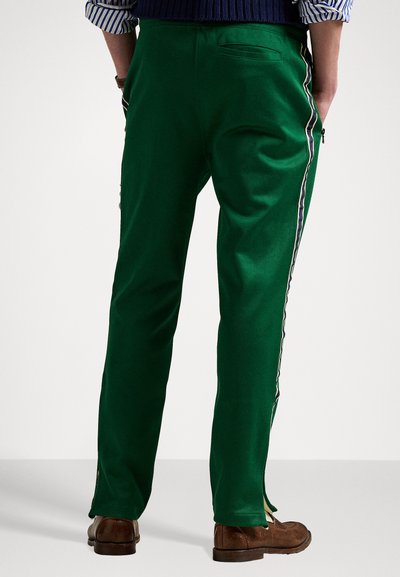 Pantaloni verzi cu o textură netedă, prezentând dungi laterale și un buzunar cu fermoar. Mânşile au deschideri, fiind asortați cu pantofi maro.