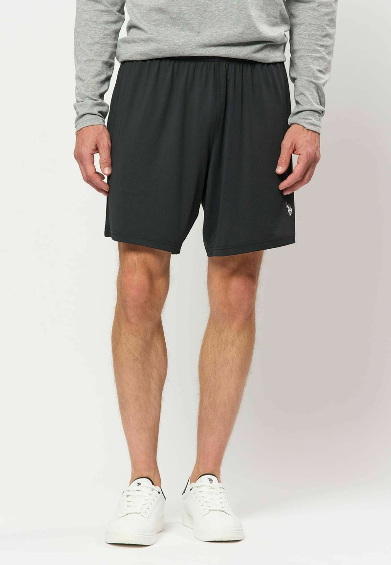 Homme portant un short noir à taille élastique, un t-shirt gris à manches longues et des baskets blanches, debout devant un fond uni.