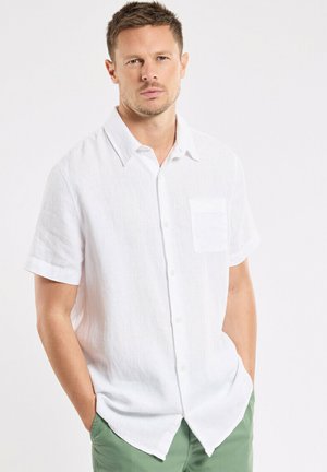Homme portant une chemise blanche à manches courtes boutonnée et un pantalon vert, debout les mains dans les poches devant un fond clair uni.