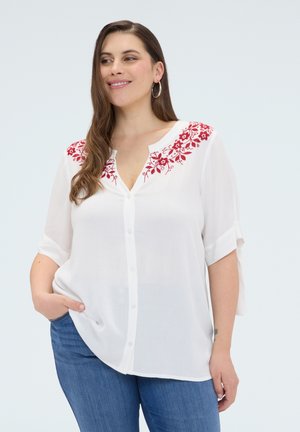 Donna che indossa una camicetta bianca con bottoni, con ricamo floreale rosso sulle spalle e jeans blu, in piedi con una mano in tasca.