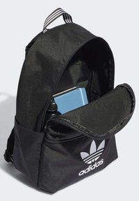 Μαύρος υφασμάτινος σακίδιο με φερμουάρ, πλάγιες τσέπες και λευκό λογότυπο Adidas. Περιέχει ένα μπλε τετράδιο και ένα μπουκάλι νερού μέσα.