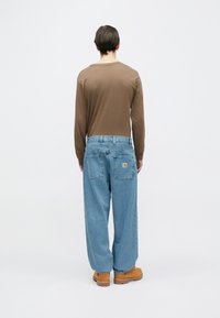 Camisa marrón de manga larga, vaqueros holgados azul claro con dos bolsillos traseros y calzado marrón claro. Diseño simple, ajuste relajado y herrajes mínimos.