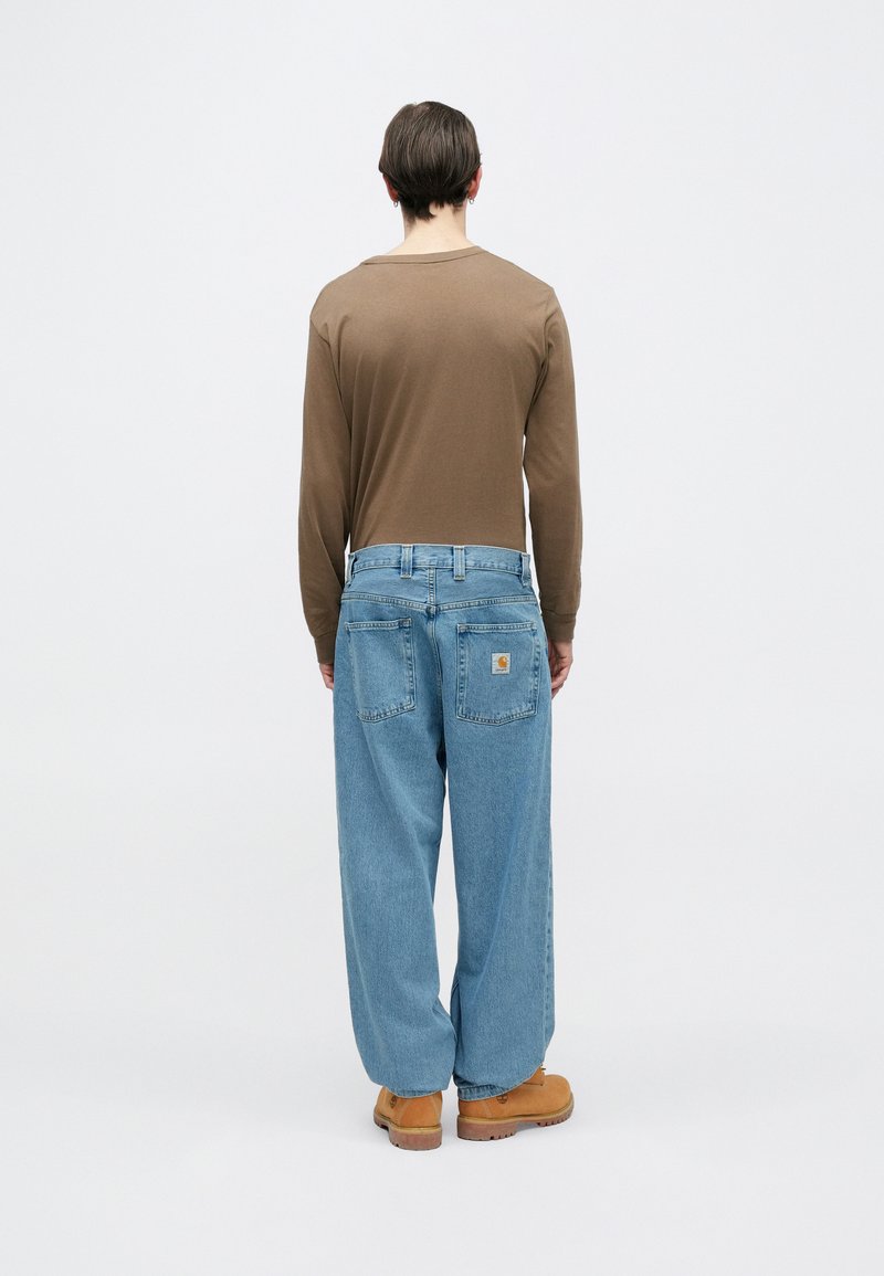 Camisa marrón de manga larga, vaqueros holgados azul claro con dos bolsillos traseros y calzado marrón claro. Diseño simple, ajuste relajado y herrajes mínimos.