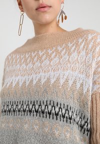Maglione beige lavorato a maglia con motivi geometrici in bianco, grigio e nero. Presenta un collo rotondo e un tessuto morbido e testurizzato.