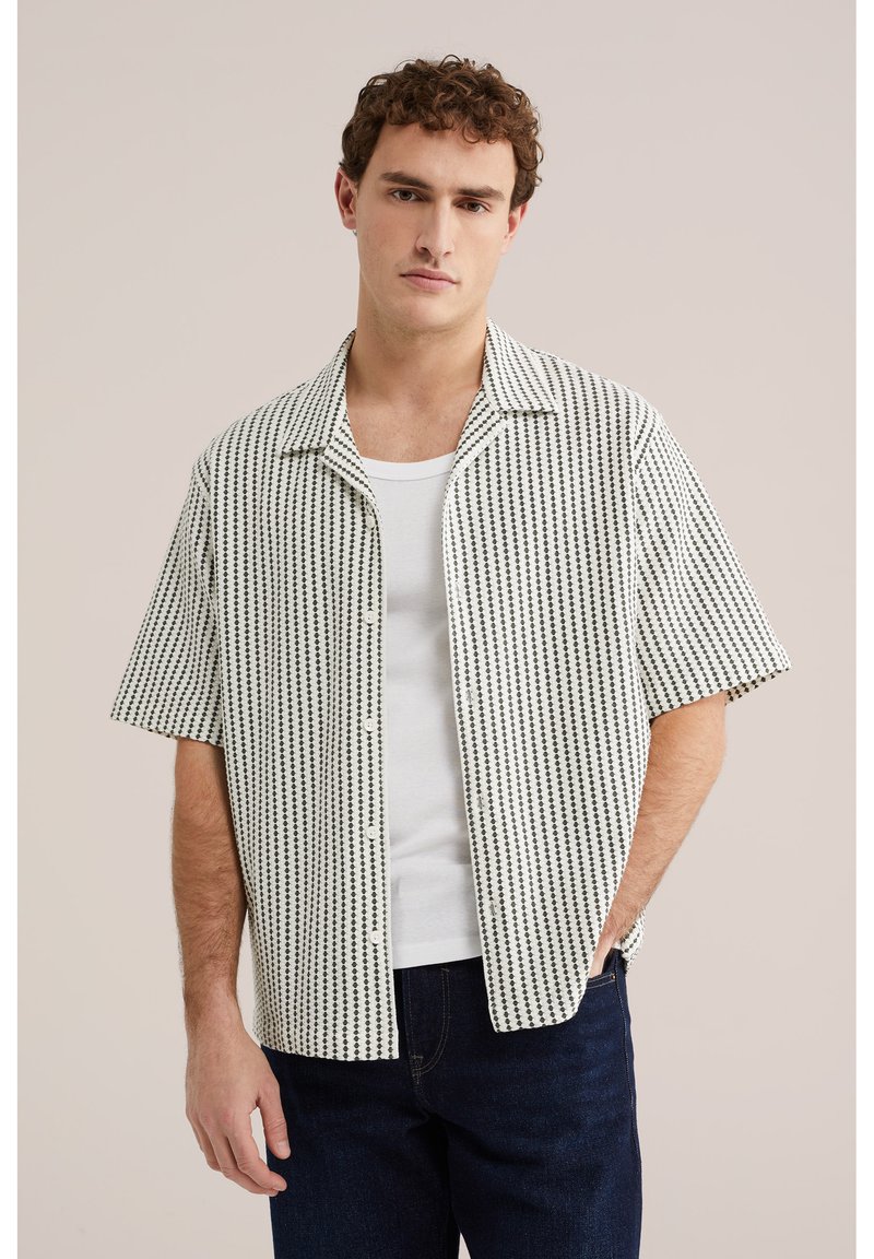 Mladić u bijeloj košulji s uzorkom i kratkim rukavima preko bijele majice i tamnoplavih traperica, stoji s jednom rukom u džepu.