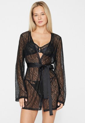Femme blonde portant une robe en dentelle noire transparente avec une ceinture en satin sur une lingerie noire assortie en dentelle, debout devant un fond clair uni.