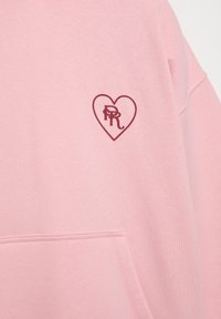 Ružová mikina s jemnou textúrou, s vínovočerveným srdcom obsahujúcim iniciály "P" a "R". Má predné vrecko.