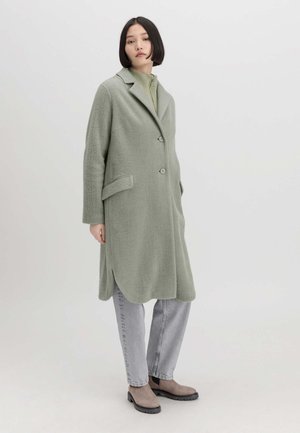 Cappotto lungo di un verde chiaro in tessuto morbido con colletto a punta, due tasche frontali e chiusura con un solo bottone. Abbinato a jeans grigi e stivali.