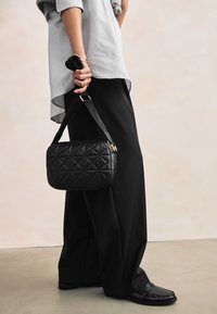 Sort quiltet skuldertaske med guldfarvet hardware, båret af en person iført en stribet skjorte og løse sorte bukser på en neutral baggrund.