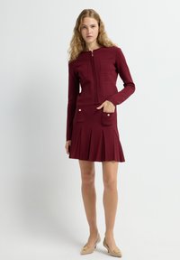 Vestido burdeos con una tela texturizada, corsé ajustado y falda acampanada con plisados; cuenta con dos bolsillos frontales con botones dorados.