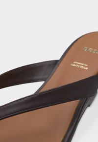 Καφέ δερμάτινο σανδάλι με δύο χοντρές λουρίδες που διασταυρώνονται πάνω από το πόδι και ταμπά πάτο με σήμανση σε χρυσό τύπωμα.