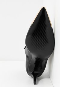 Czarne szpilki z ostrym noskiem o błyszczącym wykończeniu, z teksturowaną podeszwą, smukłym kształtem i złotym akcentem na czubku.