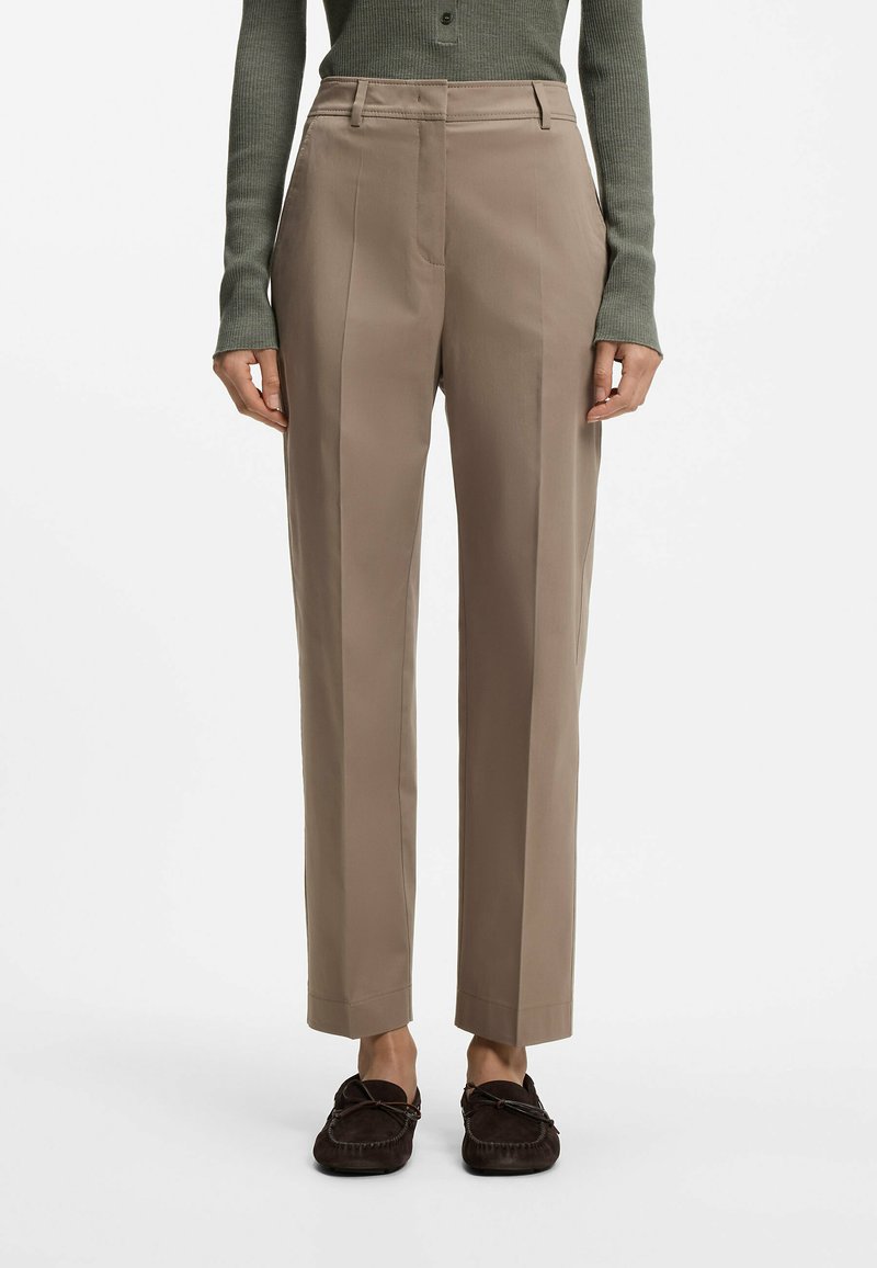 Femme portant un pantalon beige droit, des mocassins marron foncé et un haut à manches longues vert olive, debout devant un fond blanc.
