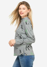 Szara koszula z długim rękawem z czarnymi i zielonymi napisami, z bocznymi rozcięciami i lekko podniesionym kołnierzem, w połączeniu z niebieskimi dżinsami.