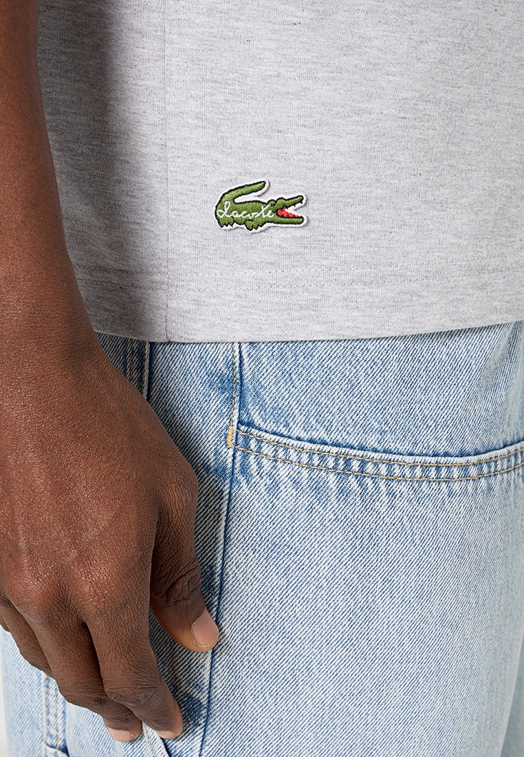 Graues Baumwoll-T-Shirt mit einem grünen gestickten Krokodil-Logo am Saum, kombiniert mit hellblauen Jeans.