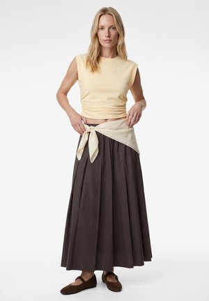 Donna che indossa un top corto beige senza maniche, gonna lunga plissettata marrone scuro, sciarpa beige annodata in vita e ballerine marroni, in piedi su sfondo bianco.