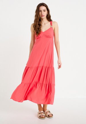 Vrouw met lang golvend haar, gekleed in een koraalroze mouwloze maxi-jurk met decoratieve broche op de schouder en beige sandalen, staand.