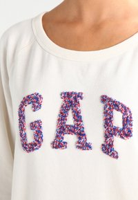 Cremefarbener Sweatshirt mit weitem Ausschnitt, der erhöhte, mehrfarbige "GAP"-Schriftzüge in roten, blauen und weißen strukturierten Fäden zeigt.
